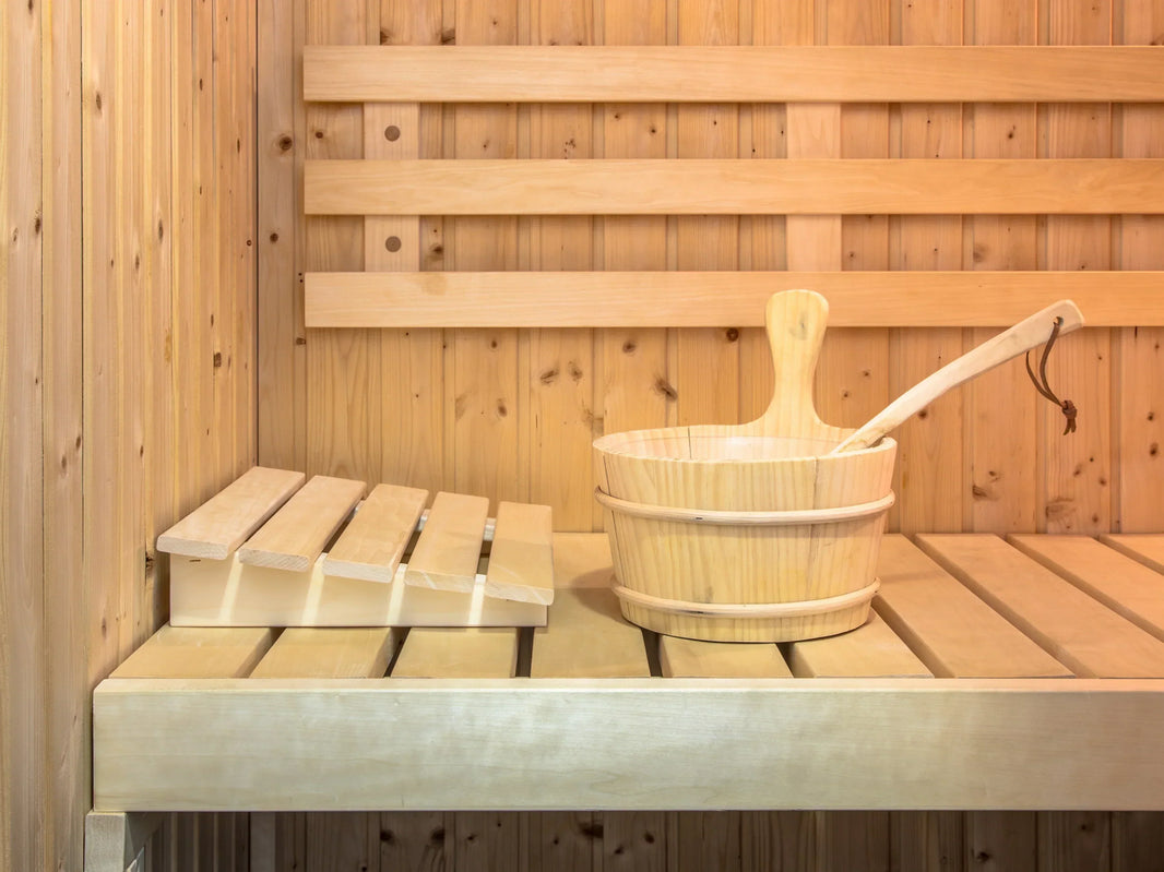 Traditional wood sauna interior with a cedar bench, headrest, and a wooden bucket and ladle used for pouring water over hot rocks.