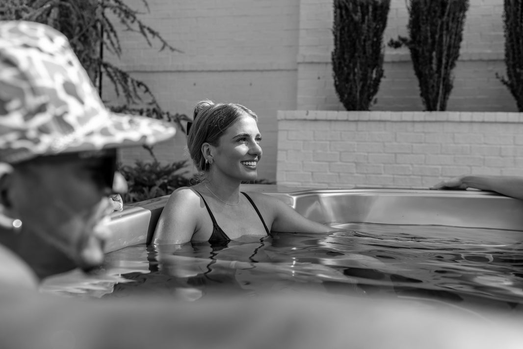Woman smiling while cold plunging outdoors next to a friend in a camo bucket hat