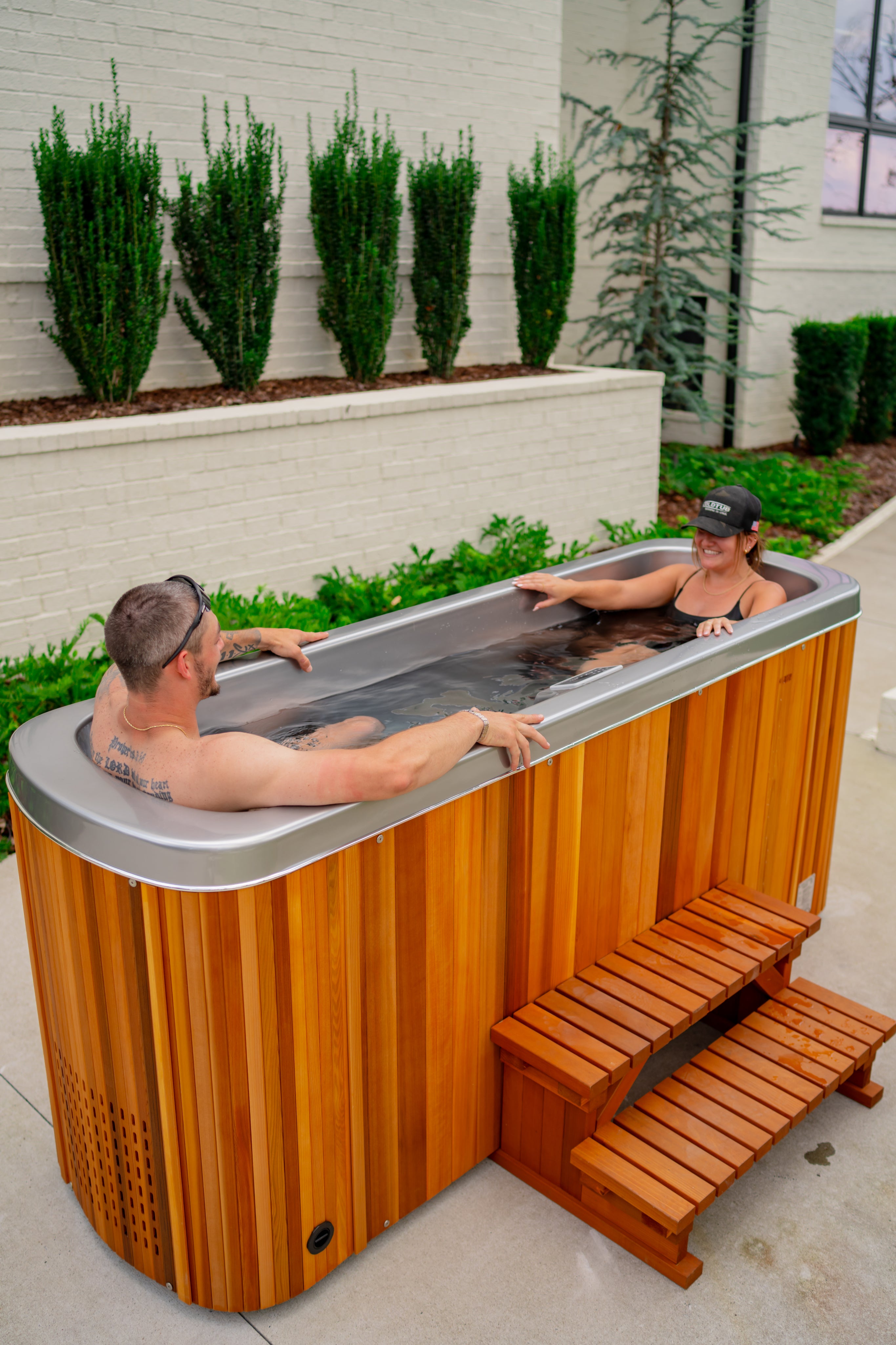 Two people are sitting and relaxing in a wooden outdoor hot tub near a white brick wall with green shrubs.