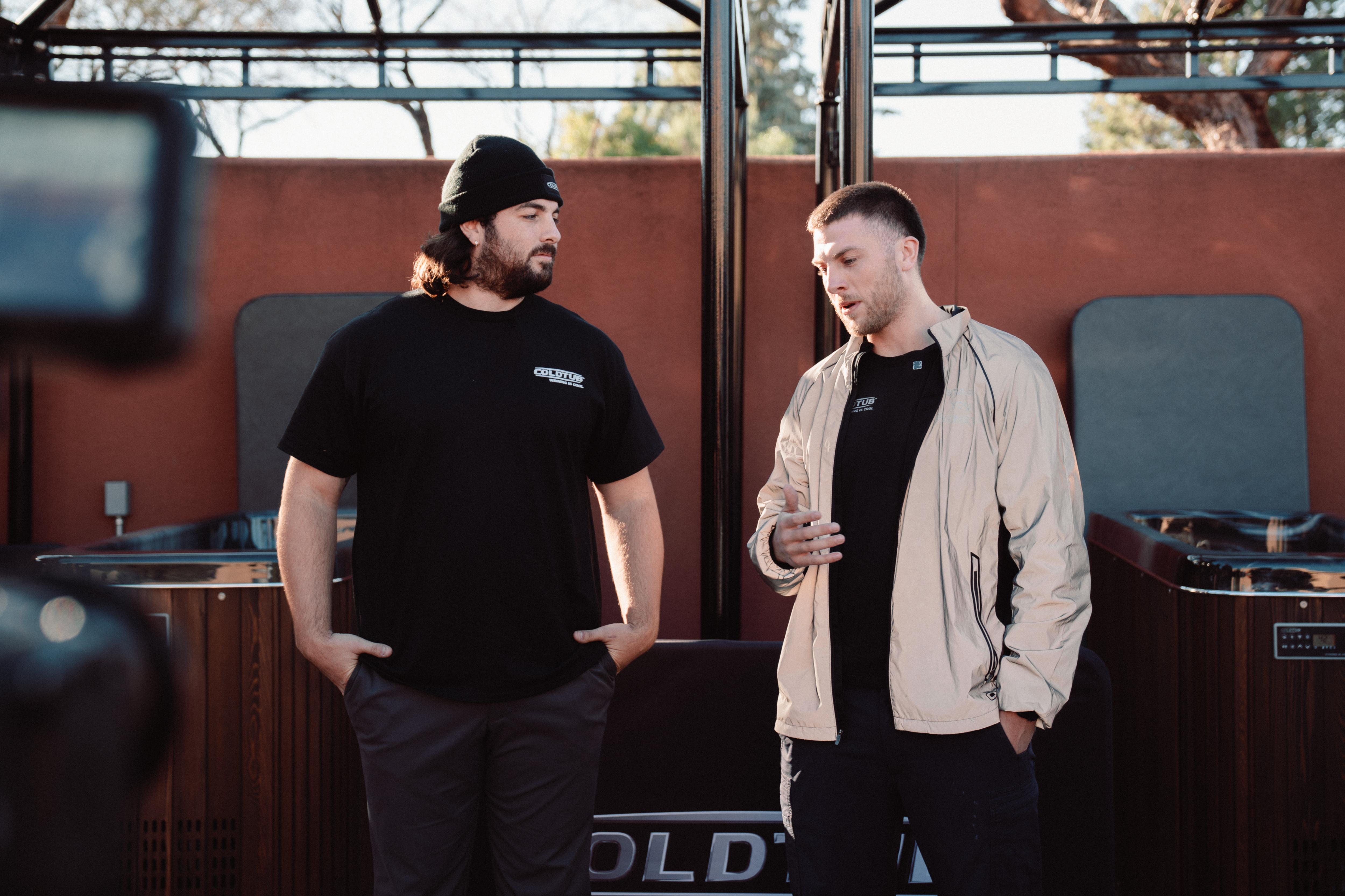 Two men stand outdoors in conversation, one wearing a black beanie and t-shirt, the other in a light jacket, with spa equipment visible in the background.
