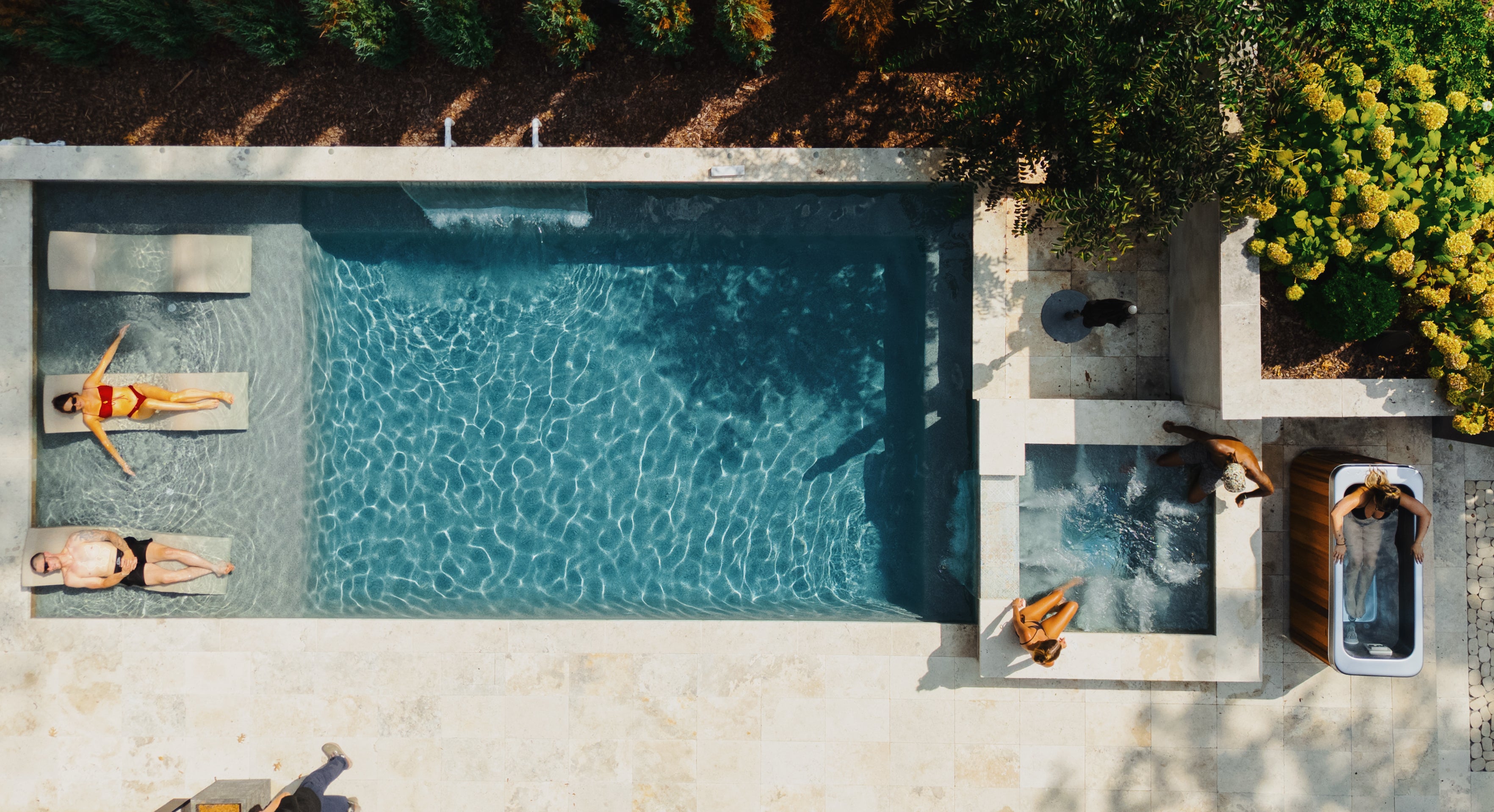 Aerial view of a rectangular pool with two people lounging on floats, two people in a hot tub, and a dog nearby on a stone patio surrounded by greenery.