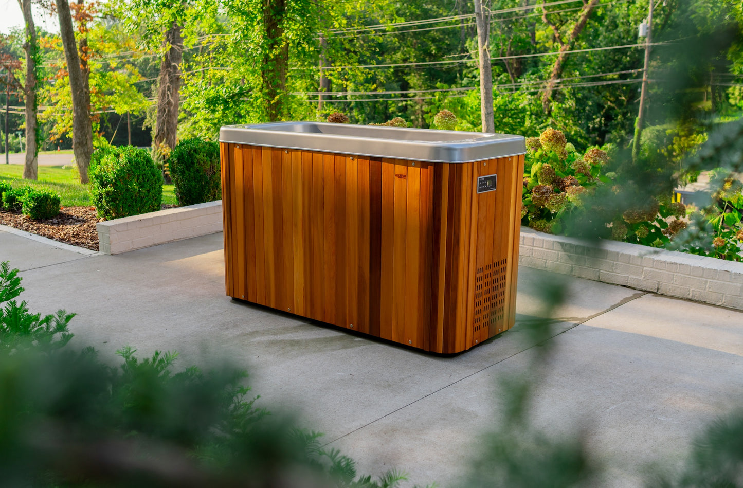 A wooden cold plunge tub with a stainless steel rim is placed on a concrete patio, surrounded by greenery and trees.