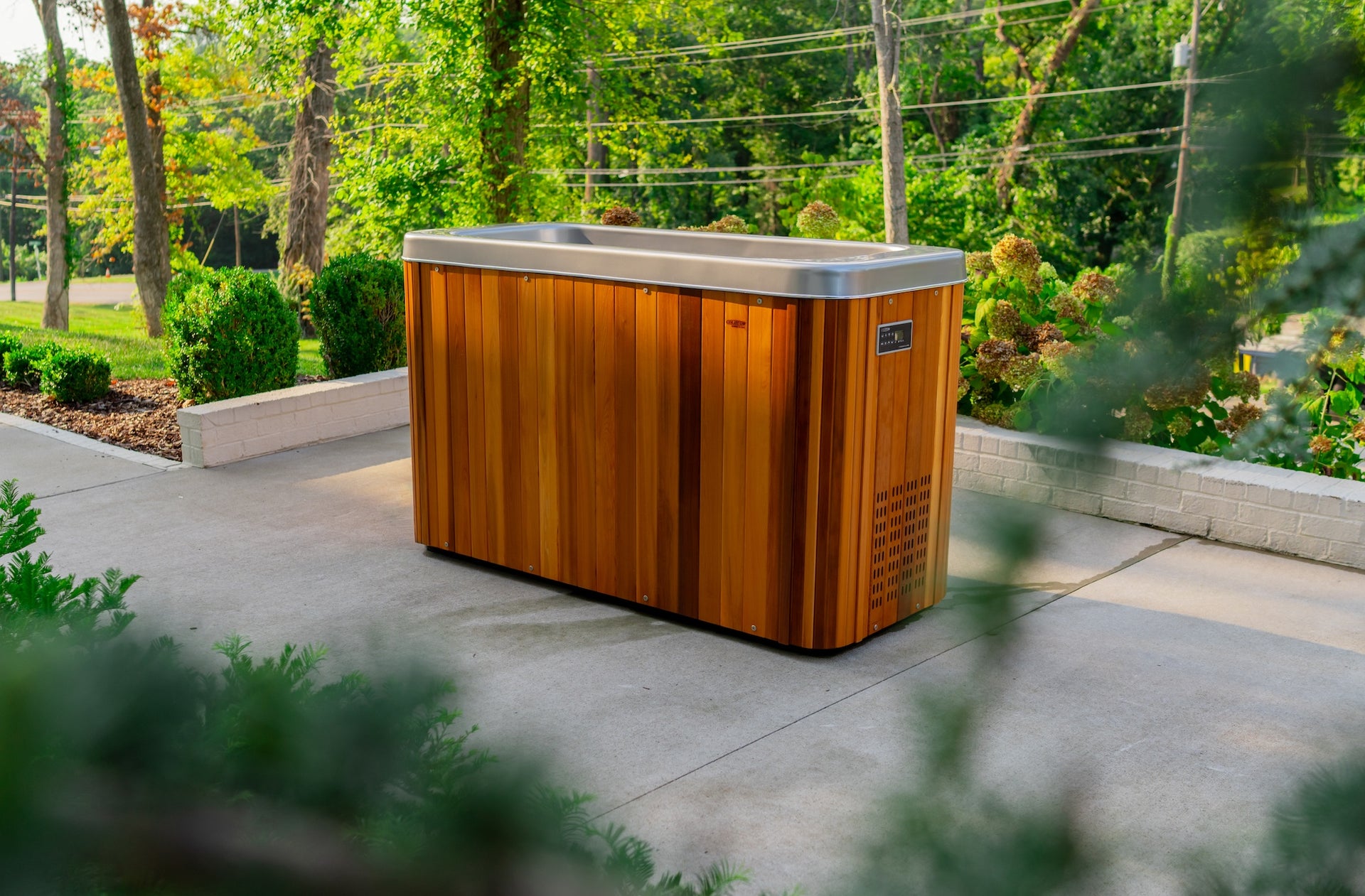 A wooden cold plunge tub with a stainless steel rim is placed on a concrete patio, surrounded by greenery and trees.