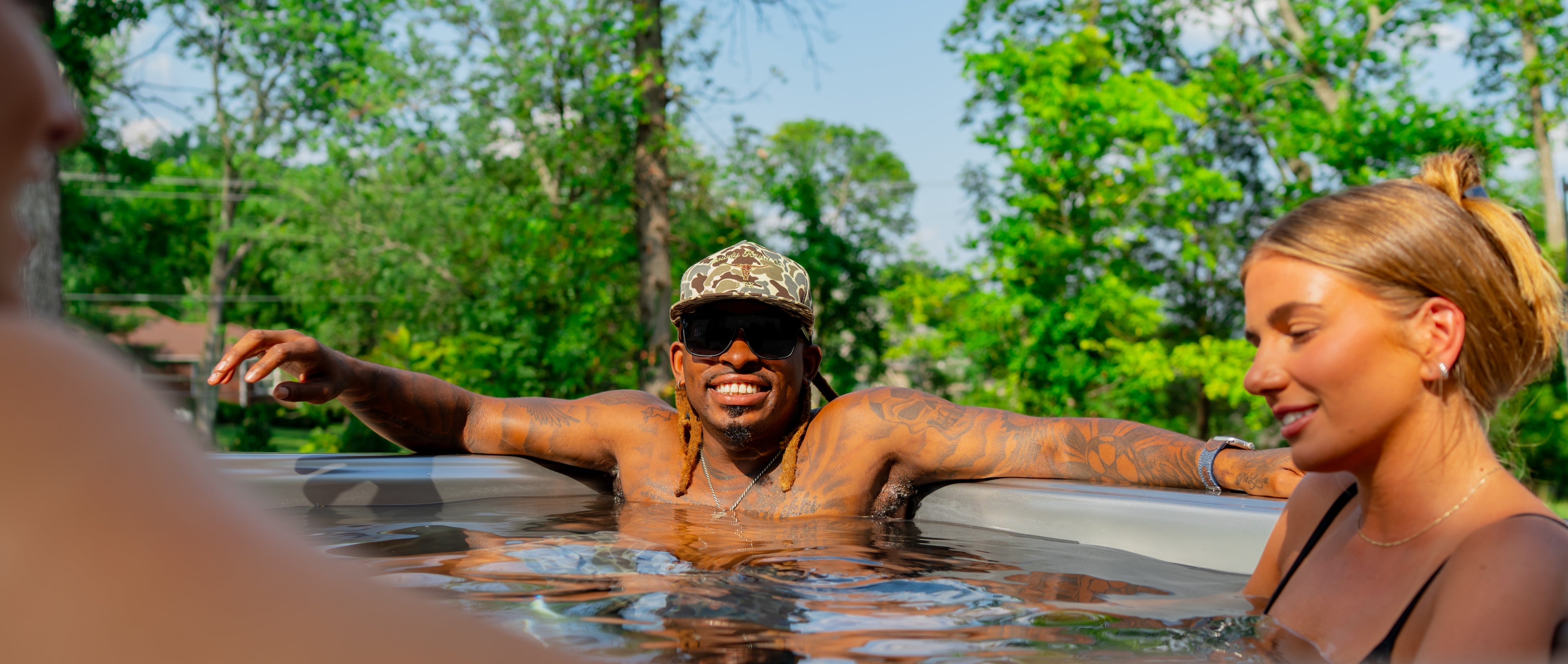 Three people relax in a hot tub outdoors on a sunny day; one man in sunglasses and a cap sits in the center, smiling, with trees in the background.