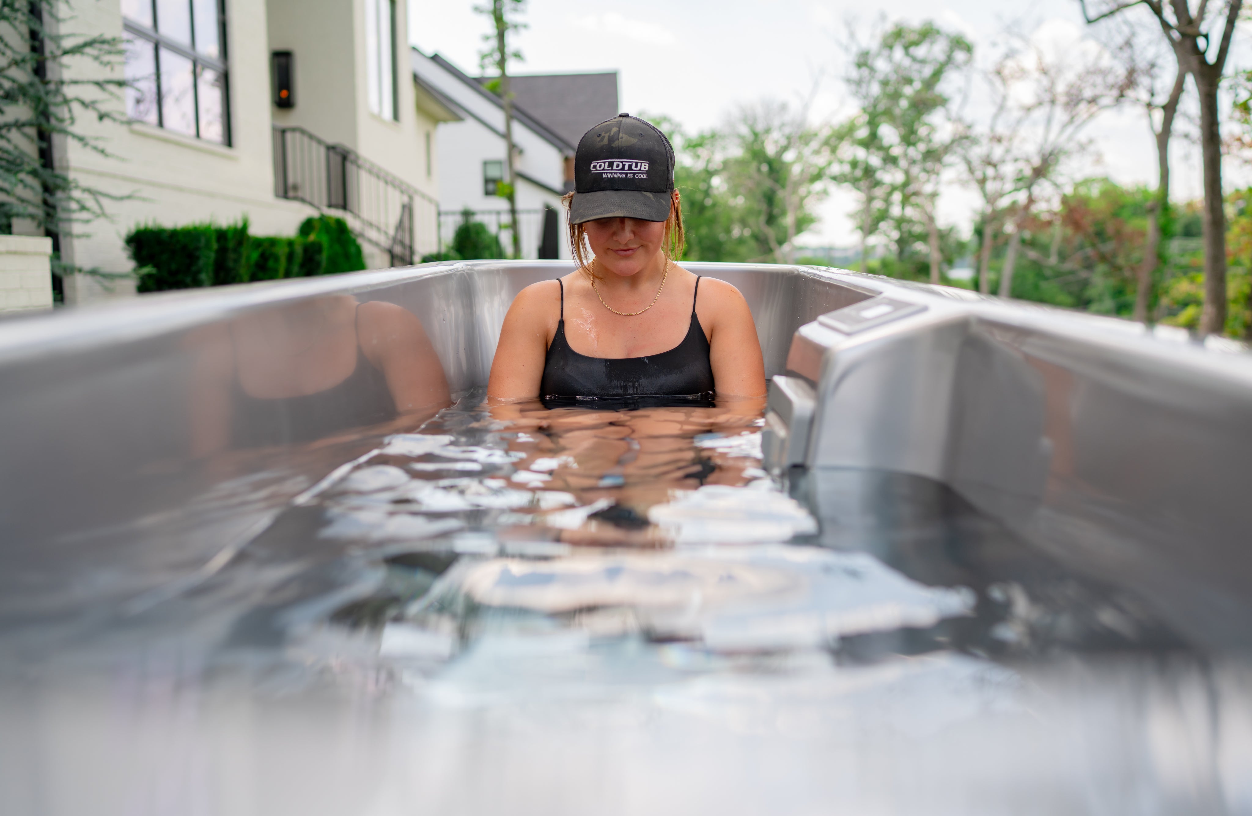A person wearing a black tank top and black hat sits in an outdoor metal tub filled with water, partially submerged, with houses and trees in the background.