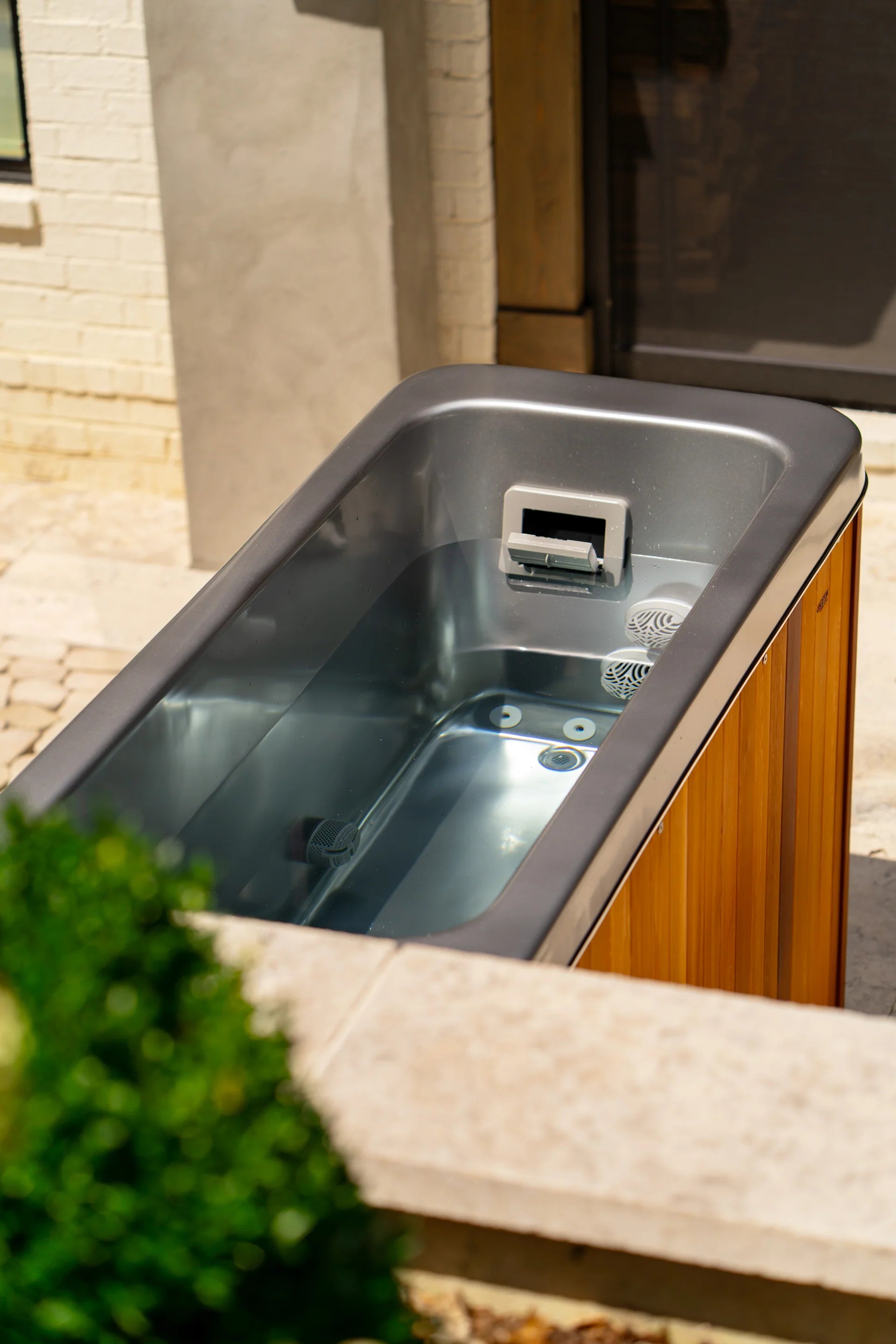 A modern outdoor soaking tub with clear water, gray interior, and wood-paneled exterior, positioned on a stone patio near a brick wall.