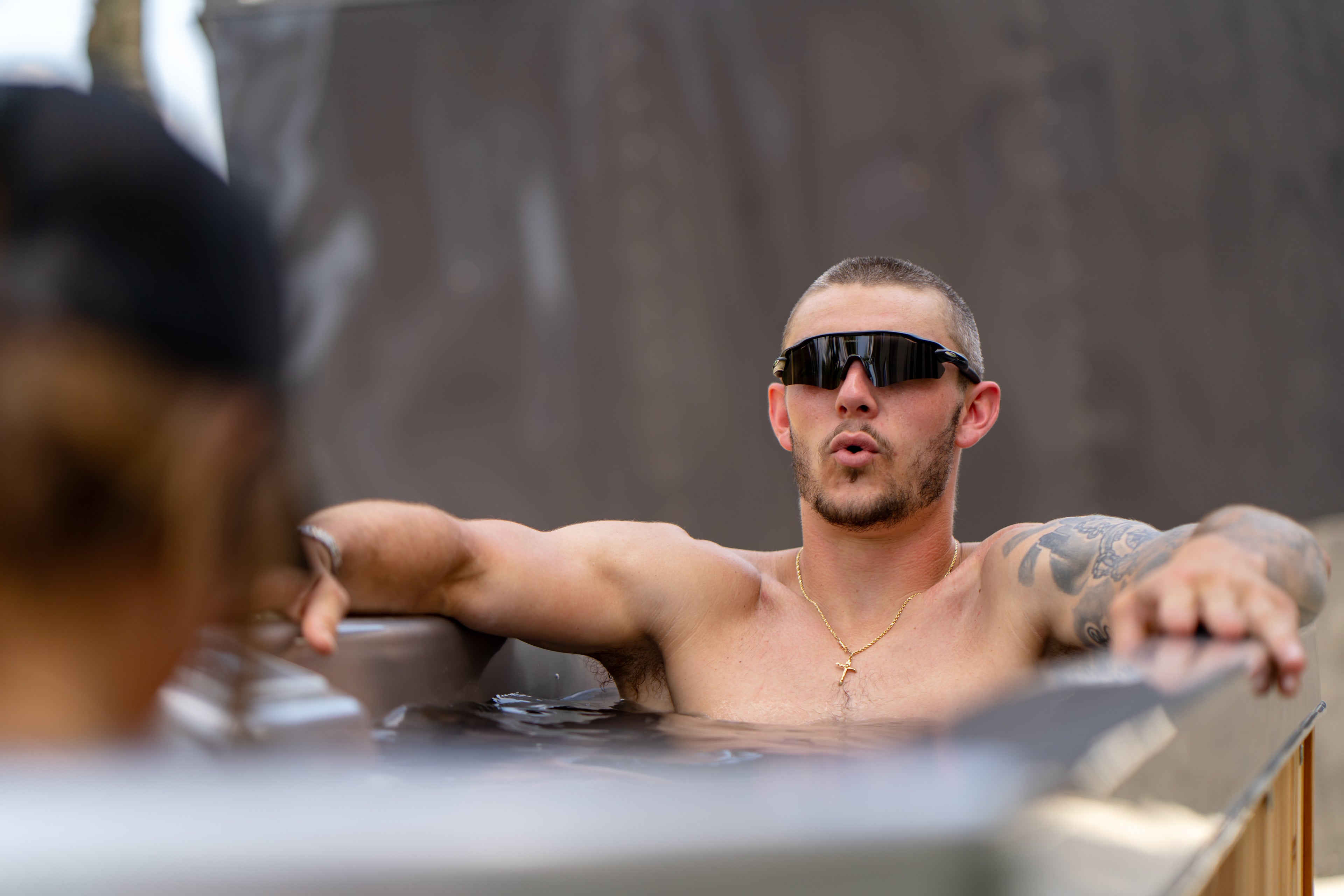 A shirtless man wearing dark sunglasses relaxes in a hot tub, arms resting on the sides, with another person partially visible in the foreground.