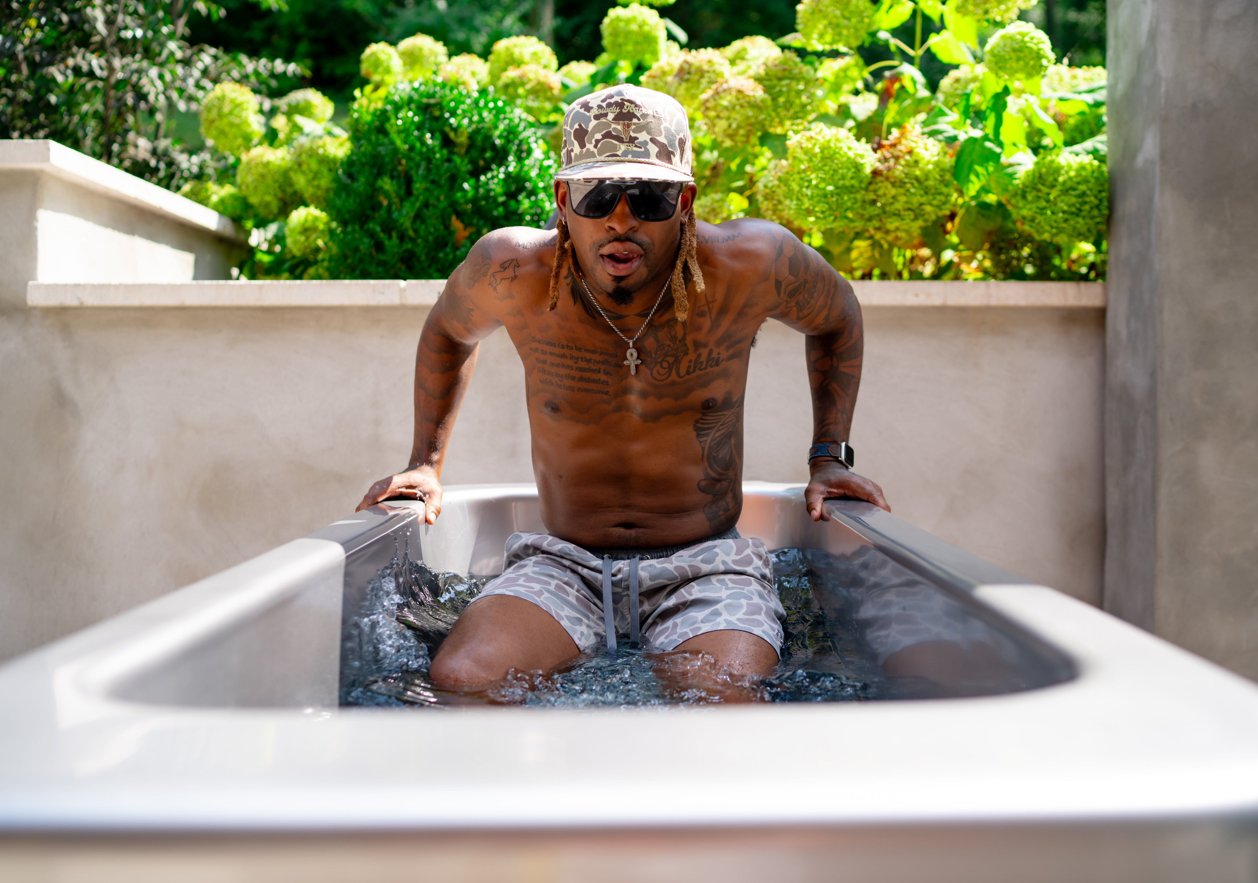 A man wearing sunglasses, a hat, and patterned swim trunks sits in an outdoor metal tub filled with water, surrounded by greenery.