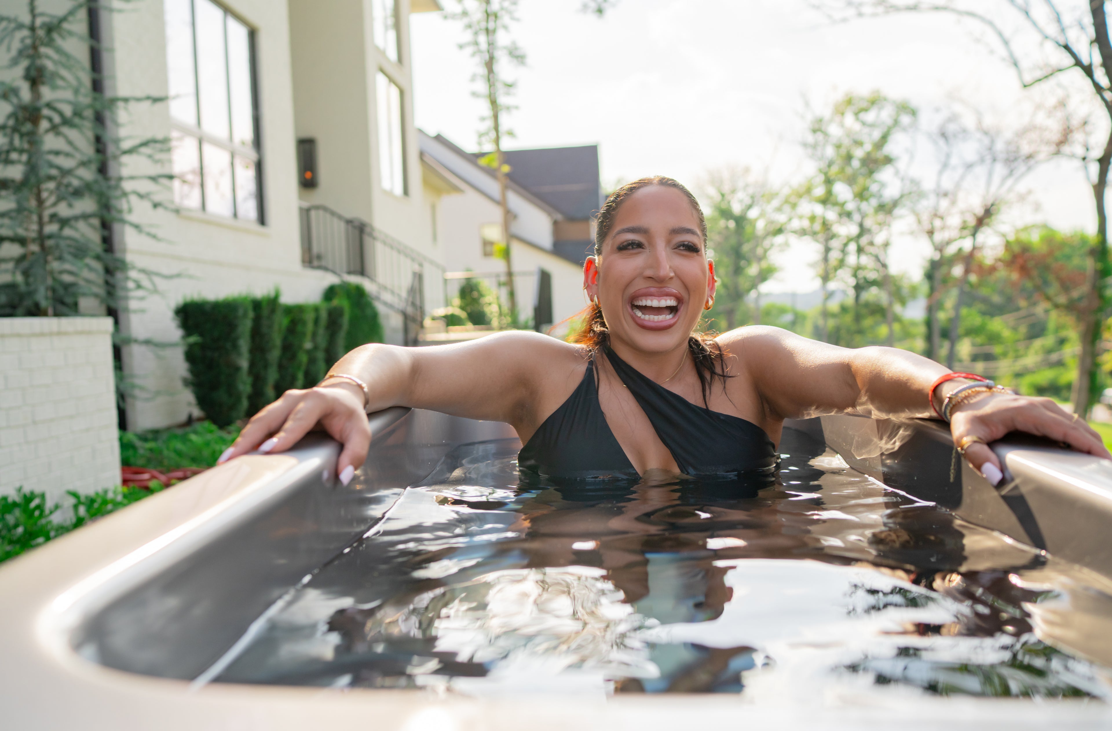 Woman in a black swimsuit smiles while sitting in an outdoor tub filled with water, with houses and trees visible in the background.